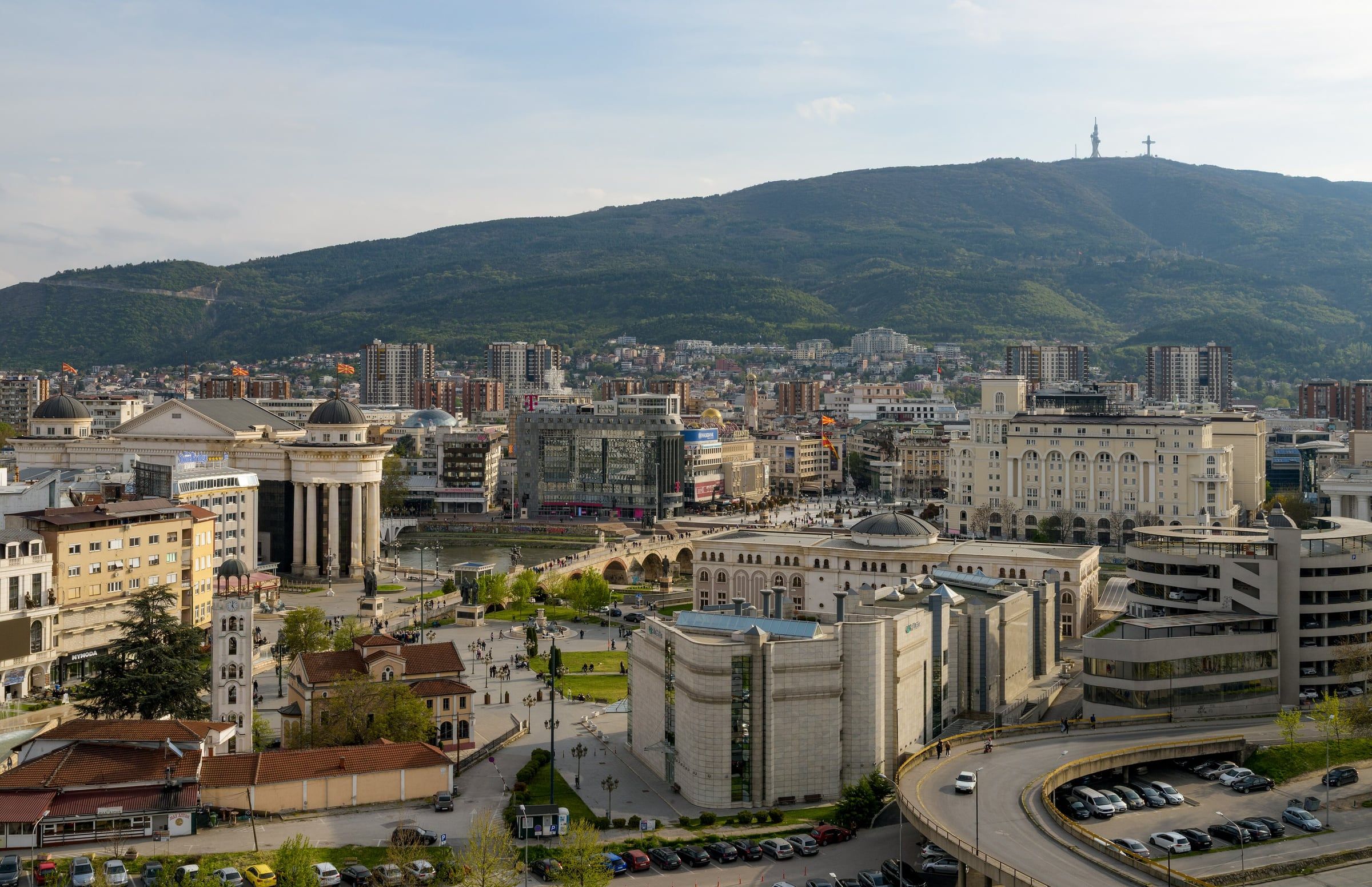 Aerial view of a city centre with an arched stone bridge over a river, domed buildings and a wooded mountain behind.