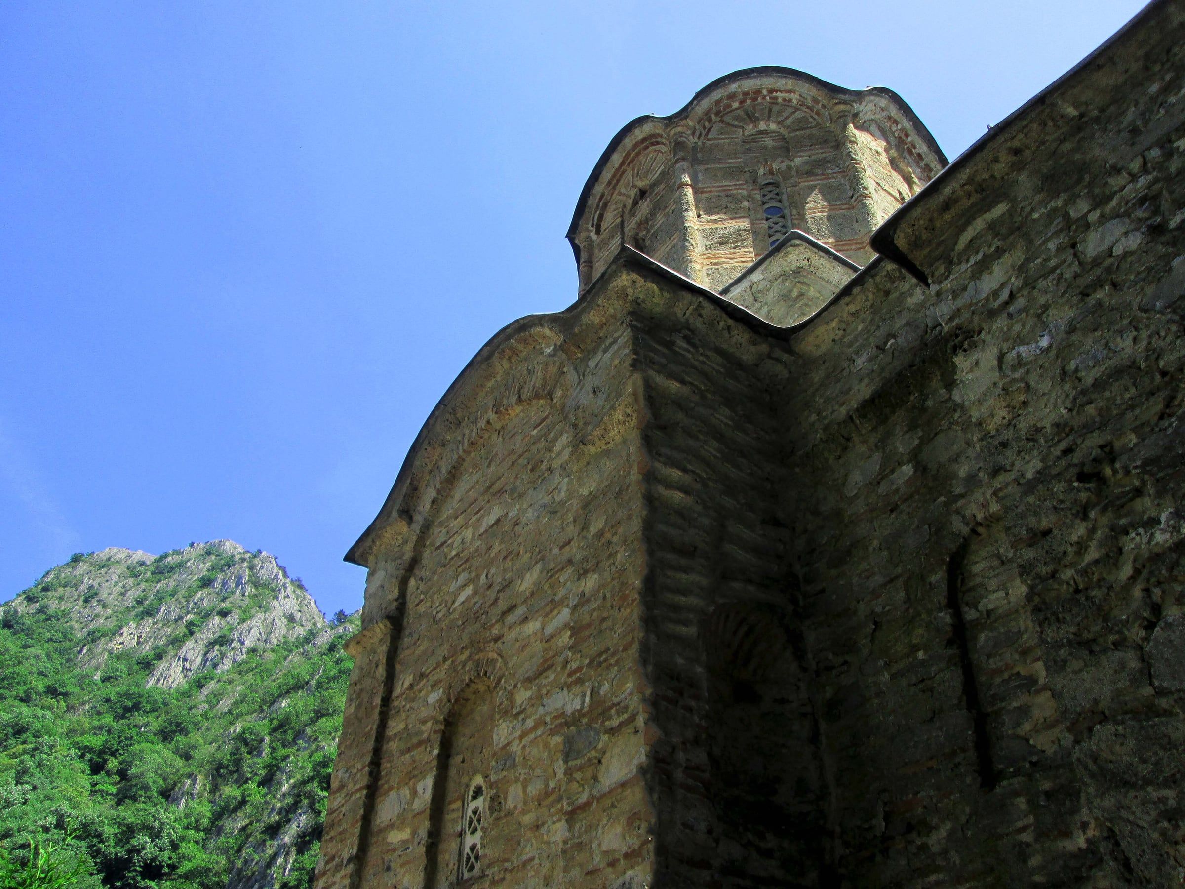 Low angle view of a weathered stone church with a small dome and a rocky cliff behind, under a clear blue sky.